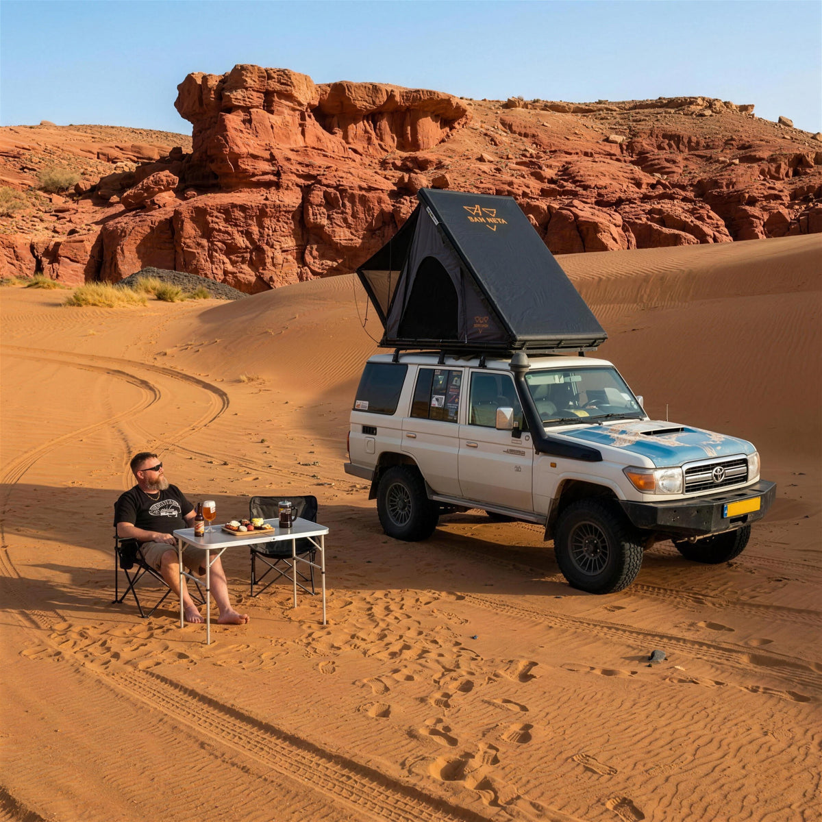 Camper at a camping table next to a 4x4 with the LCM Offroad Dune hardshell rooftop tent open in the desert